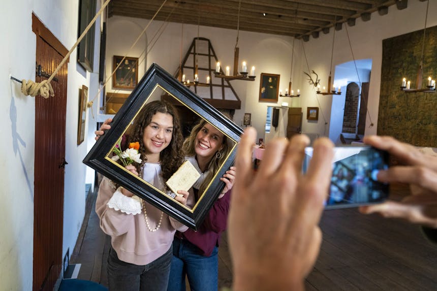 Twee vrouwelijke bezoekers houden een antiek uitziende lijst vast en kijken er lachend doorheen terwijl iemand anders op een smartphone een foto van ze maakt.
