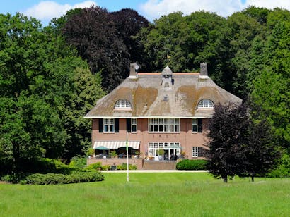 Het achteraanzicht van huis De Ehze, omgeven door bomen, met een groot gazon op de voorgrond.