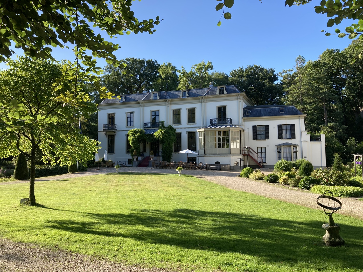 Huis te Eerbeek met serre en terras, op de voorgrond een gazon met tuinvazen en een zonnewijzer.