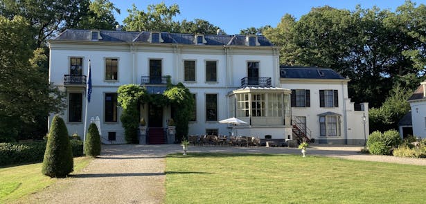 Huis te Eerbeek met serre en terras, op de voorgrond een gazon met tuinvazen en in vorm gesnoeide struiken.