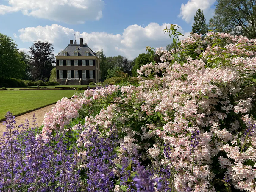Huis Verwolde, zichtbaar achter een groen gazon, met kleurrijke roze en blauw-paarse bloemen langs een tuinpad op de voorgrond.