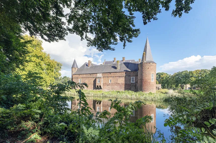 Achterzijde van kasteel Hernen, een middeleeuws bakstenen kasteel met ronde torens en puntdaken, omgeven door groen en gereflecteerd in de omliggende slotgracht.
