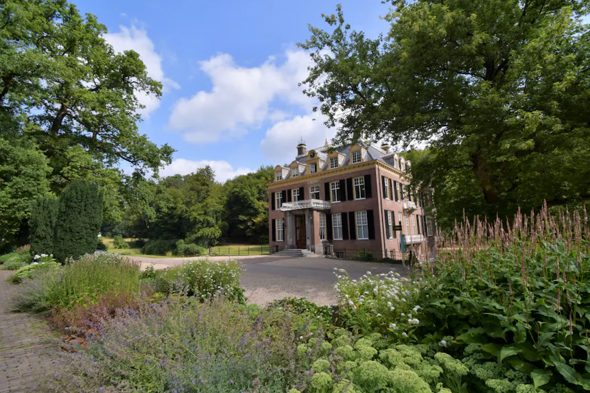 Landhuis Zypendaal, een statig historisch huis met een symmetrische gevel van baksteen, witte kozijnen, donkere luiken en een kleine trap bij de entree. Op de voorgrond liggen verzorgde bloemperken met groene en bloeiende planten, terwijl het landhuis wordt omringd door een parkachtig landschap met hoge bomen.