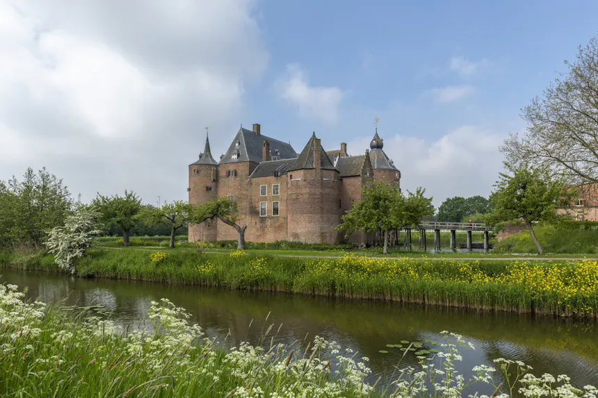 Kasteel Ammersoyen: een groot bakstenen kasteel met meerdere ronde hoektorens en puntige leien daken, kleine ramen met luiken en een houten toegangsbrug. Het kasteel is zichtbaar vanaf een groene, begroeide waterkant, onder een lichtblauwe lucht met verspreide witte wolken.