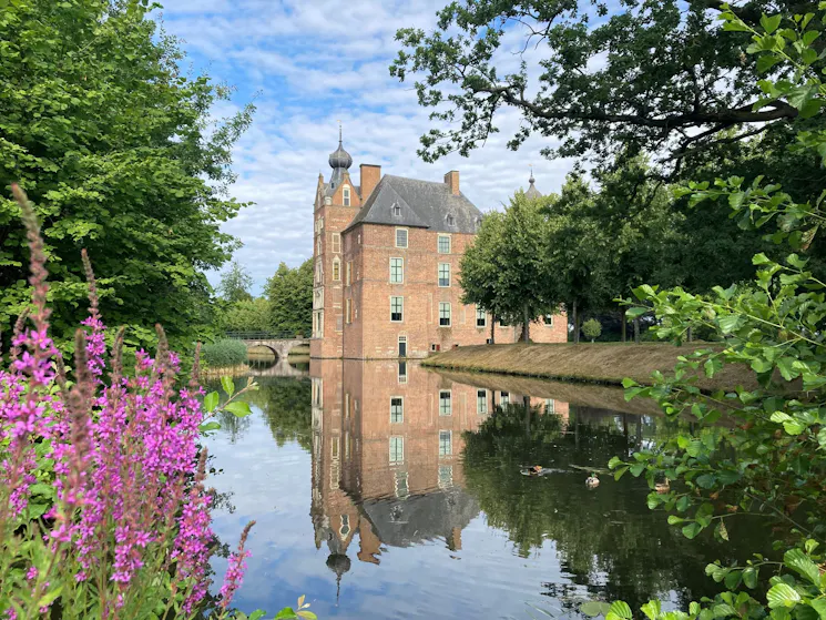 Zijaanzicht van Kasteel Cannenburch, een groot bakstenen gebouw met een toren met uivormige spits en leien daken, gelegen aan een gracht en omringd door loofbomen. Op de voorgrond staan paarse bloemen langs de oever, terwijl op de achtergrond een stenen brug zichtbaar is. Het kasteel wordt duidelijk weerspiegeld in het water.