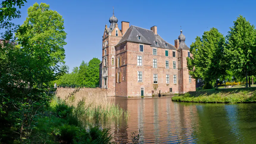 Zijaanzicht van Kasteel Cannenburch in de zon, een groot bakstenen kasteel met twee torens met uivormige spitsen en leien daken. De hoofdingang en de stenen toegangsbrug zijn zichtbaar. Het kasteel ligt aan een gracht met rietkragen, omringd door groene loofbomen.