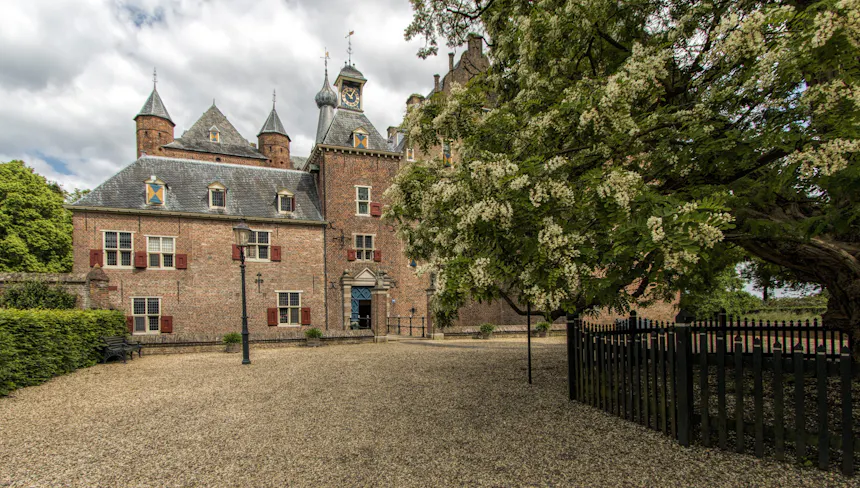 Kasteel Doorwerth, een bakstenen kasteel met glas-in-loodramen voorzien van luiken, meerdere torens en een dak van leisteen, gezien vanaf een open grindplein. In de voorgevel bevindt zich centraal een toegangspoort met omlijsting. Rechts op de afbeelding staat een grote acaciaboom met witte bloesem.