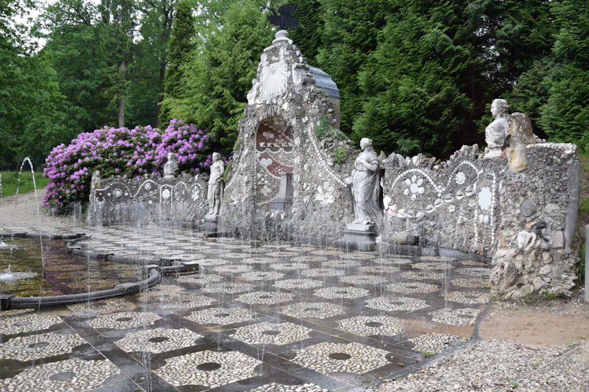 Afbeelding van de fonteinpartij De Bedriegertjes in het park van Kasteel Rosendael. Verborgen 18e-eeuwse fonteinen spuiten onverwacht water uit de bestrating. Op de achtergrond zijn een muur met schelpen, tuinbeelden en groen zichtbaar.