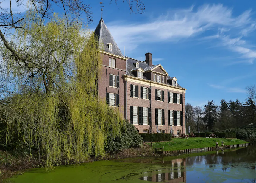 Huis Verwolde, een historisch bakstenen landhuis met meerdere verdiepingen, groene luiken, een zadeldak met dakkapellen en een toren, gelegen aan een gracht en omgeven door parkachtig groen.