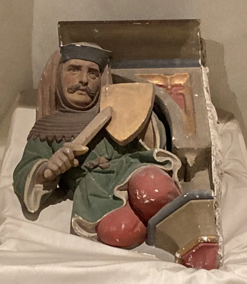 Historisch stenen bouwfragment uit kasteel Ammersoyen, tijdelijk verwijderd voor restauratie. Afgebeeld is een mannelijke figuur in middeleeuwse kleding, met snor en hoed, die een klein schild en een zwaard vasthoudt. De figuur is beschilderd in groen, rood en goud.