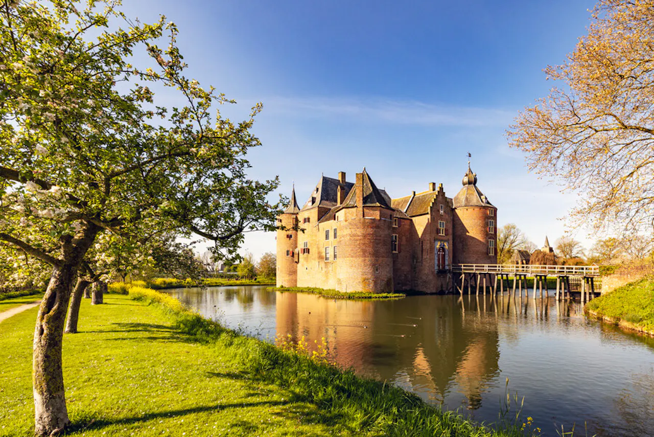 Voor- en zijaanzicht van kasteel Ammersoyen, omgeven door een brede slotgracht, badend in het zonlicht en onder een strakblauwe lucht. Drie van de vier imposante, ronde hoektorens zijn zichtbaar, samen met de tussenliggende vleugels. Een houten brug over de slotgracht leidt naar het poortgebouw.