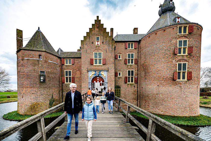 Vooraanzicht van kasteel Ammersoyen met een trapgevel en imposante ronde torens, waarvan één een uivormige torenspits heeft. Boven de toegangspoort prijkt een wapen. Bezoekers lopen over een houten brug die over de slotgracht voert.